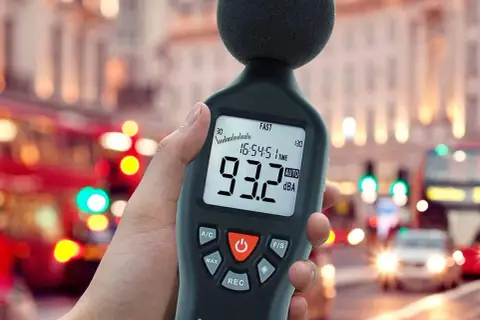 Medidor de sonido en mano, mostrando 93.2 dBA en una calle ruidosa, midiendo la potencia acústica del tráfico urbano.