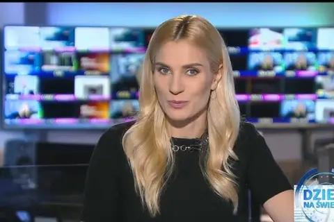 Blond dziennikarka TVN w czarnej bluzce z naszyjnikiem, na tle monitorów.