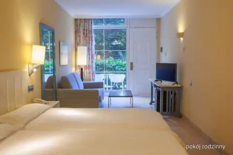 Pokój rodzinny w hotelu IFA Altamarena: łóżko, sofa, TV i wyjście na taras z widokiem na zieleń.