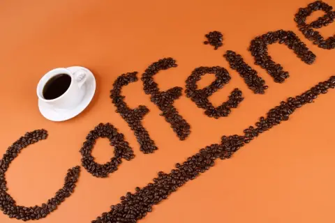 Ziarna kawy ułożone w napis "caffeine" obok filiżanki kawy. Zastanawiasz się, ile kofeiny ma łyżeczka kawy mielonej?