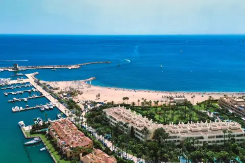 Widok na marinę pełną jachtów i piaszczystą plażę, gdzie rozciąga się złoty wybrzeże. W tle błękitne morze i horyzont.