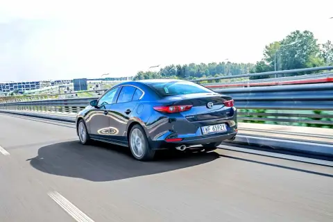 Ciemnoniebieska Mazda 3 sedan jedzie po autostradzie, odbijając światła i otoczenie.
