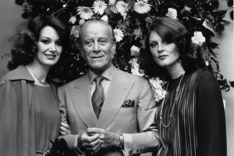 Rodzina Gucci: założyciel Gucci z dwiema kobietami na tle kwiatów.