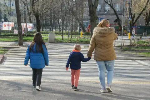 Rodzina przechodzi przez pasy. Kobieta trzyma za rękę małego chłopca, a obok idzie starsza dziewczynka. Szczególna ostrożność kiedy przechodzimy przez jezdnię.