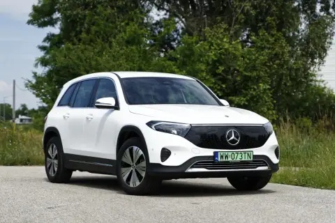 Biały Mercedes EQB na tle zieleni i nieba. Elektryczny SUV z charakterystycznym grillem i nowoczesnymi światłami.