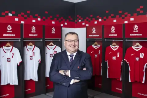 Uśmiechnięty mężczyzna w garniturze stoi przed szatnią reprezentacji Polski. Wiszą tam koszulki z nazwiskami Milika, Lewandowskiego i innych.