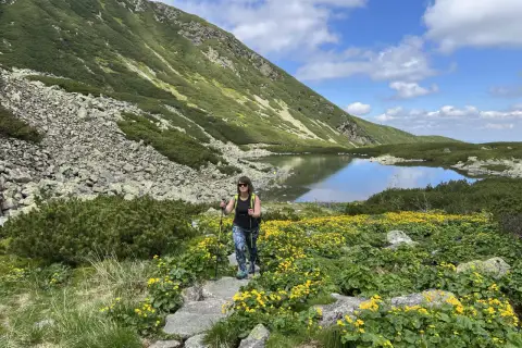 Kobieta z kijkami trekkingowymi idzie przez kwitnącą łąkę w górach. To najłatwiejszy szlak na Krzyżne, z jeziorem w tle.