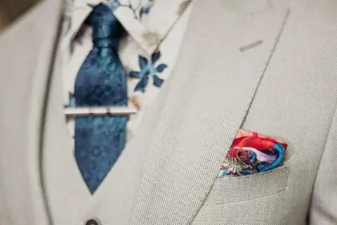 Elegancki mężczyzna w jasnej marynarce, z niebieskim wzorzystym krawatem i spinką do krawata jak nosić. W kieszeni kolorowa poszetka.