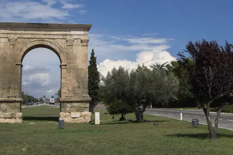 Arco romano de piedra, parte de una antigua roda de bara, flanqueado por árboles y palmeras bajo un cielo azul con nubes.