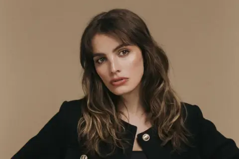 Retrato de una mujer con cabello castaño ondulado y flequillo, luciendo un blazer negro. Su mirada es intensa, transmitiendo un aire de misterio y elegancia, como si fuera la protagonista de una historia de lux pascal.
