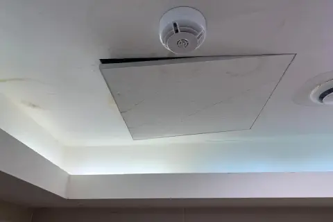 Un soffitto in cartongesso con una botola aperta, un rilevatore di fumo e macchie d'acqua. Il montaggio botola cartongesso è visibile.