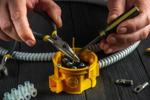 Ręce majsterkowicza wykonują wymianę puszki elektrycznej, używając szczypiec i śrubokręta do podłączenia przewodów.