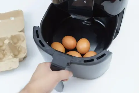 Ręka wkłada jajka do frytkownicy beztłuszczowej. Obok leży wytłaczanka. Szybkie i zdrowe jajka z airfryer.