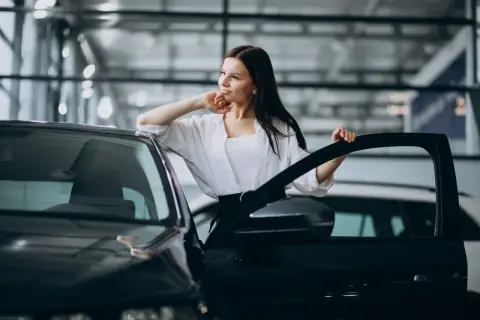 Kobieta w białej koszuli opiera się o drzwi czarnego samochodu, rozważając najem samochodu osobowego.