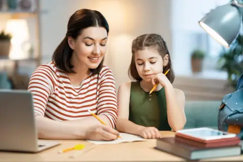 Mama pomaga córce w nauczaniu domowym podstawówka. Razem piszą w zeszycie, obok leży laptop.