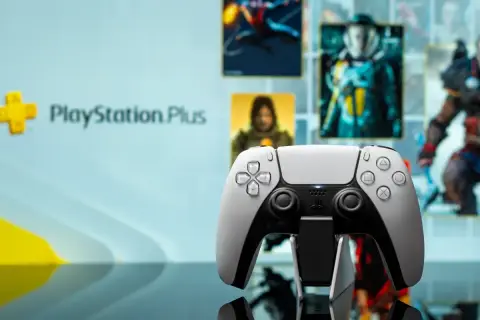 Biały kontroler DualSense na tle ekranu z logo PlayStation Plus i okładkami gier. Idealny do grania w gry z oferty PS Plus marzec.