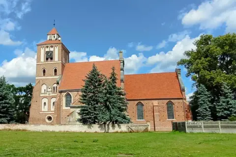 Nieszawa atrakcje: ceglany kościół z wieżą i zegarem, otoczony zielenią, pod błękitnym niebem z chmurami.