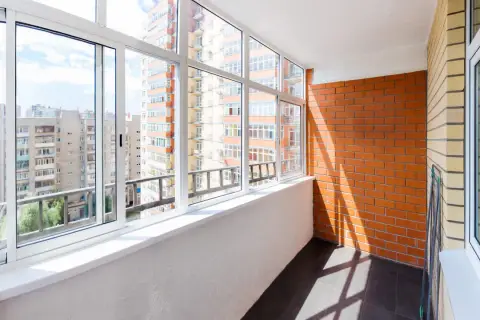 Szklana loggia z widokiem na blokowisko. Jasny balkon z białymi oknami i ceglaną ścianą.
