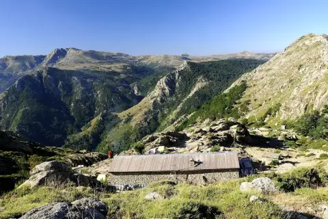 Kamienny schron na GR20 Korsyka, otoczony skalistymi zboczami i zielonymi dolinami.