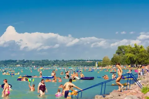 Tłum ludzi cieszy się słońcem i wodą na zatłoczonej Balaton plaży. Dzieci i dorośli pływają, relaksują się na materacach i bawią się w wodzie.