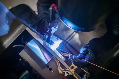 Precyzyjne spawanie TIG metalowych elementów. Ręce w rękawicach trzymają palnik i drut spawalniczy, tworząc jasny łuk.