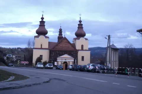 Cerkiew łemkowska z charakterystycznymi kopułami, otoczona samochodami, na tle pagórkowatego krajobrazu.