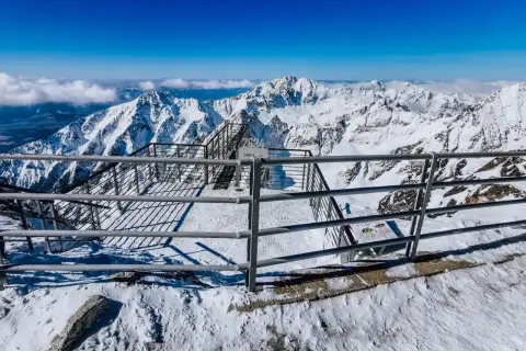 Widok z tarasu widokowego na szczycie Łomnicy. Stalowe balustrady i schody prowadzą do zapierającej dech w piersiach panoramy ośnieżonych gór.