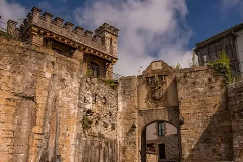 Antigua muralla y arco de entrada en Fuenterrabía, con un escudo nobiliario y un cielo azul con nubes.