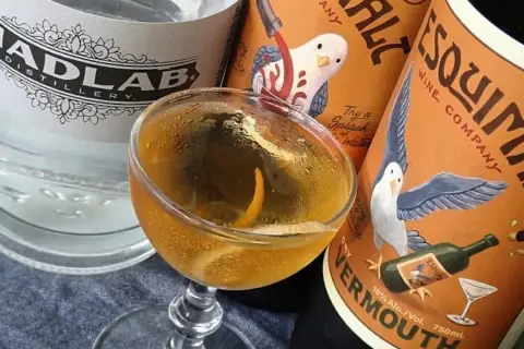 Un cocktail martini doré, garni d'un zeste d'orange, est prêt à être dégusté, entouré de bouteilles de gin et de vermouth.
