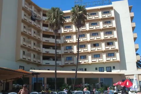 Paloma vuela frente al Hotel Royal Costa, un edificio de varios pisos con balcones y palmeras.
