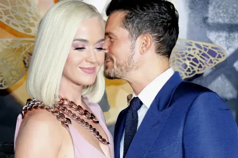 Orlando Bloom szepcze coś do ucha Katy Perry, która ma na sobie różową sukienkę i gruby, złoty naszyjnik.
