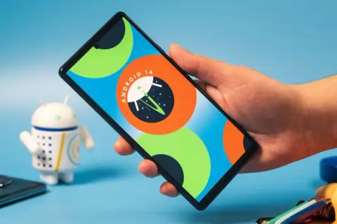Dłoń trzyma smartfon z logo Android 14. Obok stoi figurka robota.