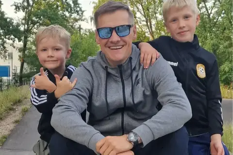 Marcin Mroczek z uśmiechem i dwoma synami, którzy go obejmują. Radosny dzień ojca i dzieci.
