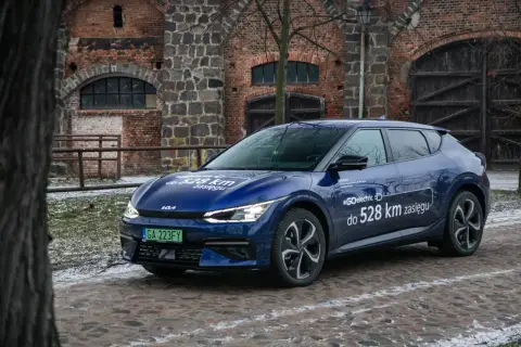 Niebieska Kia EV6 z napisem "#GOelectric D do 528 km zasięgu" stoi przed zabytkową ceglaną budowlą.