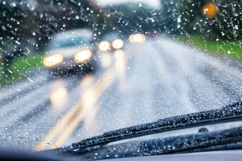 Gotas de lluvia en el parabrisas, luces borrosas de otros coches y la carretera mojada. Conducir con lluvia requiere precaución.