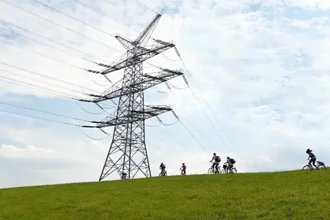 Ludzie na rowerach jadą po zielonym wzgórzu pod ogromną konstrukcją linii wysokiego napięcia.