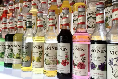 Rząd butelek syropów do kawy Monin o różnych smakach: od owocowych po kwiatowe, np. róża, lawenda, hibiskus.