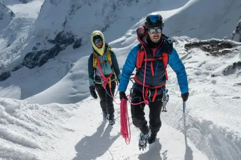 Dwóch alpinistów wspinających się po ośnieżonym zboczu, z liną i sprzętem. Zapewniają sobie asekurację lotną w trudnym terenie.