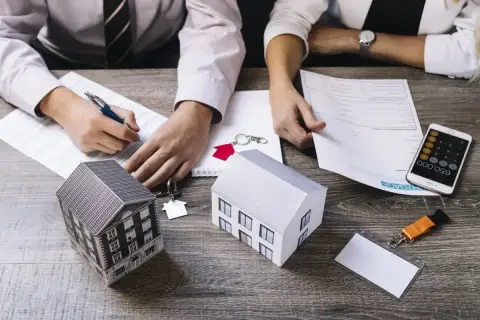 Dwie osoby analizują dokumenty, obok modele domów i klucze. To obrazuje, co to jest kredyt hipoteczny i jak go uzyskać.
