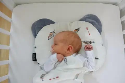 Nowy dzidziuś śpi na poduszce dla niemowlaka w kształcie misia, otulony kocykiem z piórkami.