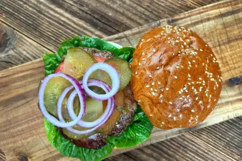 Pyszne mięso na burgery z dodatkami: sałatą, ogórkiem kiszonym i czerwoną cebulą, na desce z drewna.