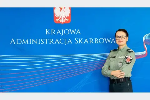 Kobieta w mundurze służby celno-skarbowej stoi przed niebieskim tłem z napisem "Krajowa Administracja Skarbowa".