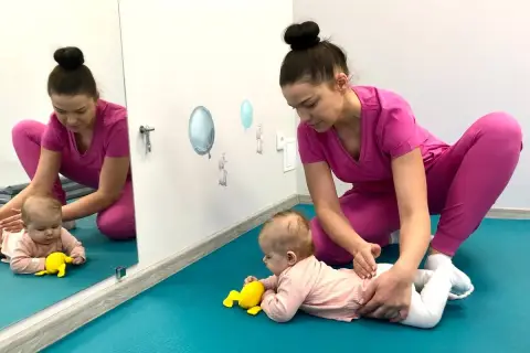 Fizjoterapeutka wspiera niemowlę na brzuchu, stosując metodę PNF. Dziecko ćwiczy podnoszenie główki, a terapeuta delikatnie koryguje jego ułożenie.