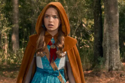 Holly Wheeler z Stranger Things w brązowym płaszczu z kapturem i niebieskiej sukience, stoi w lesie.