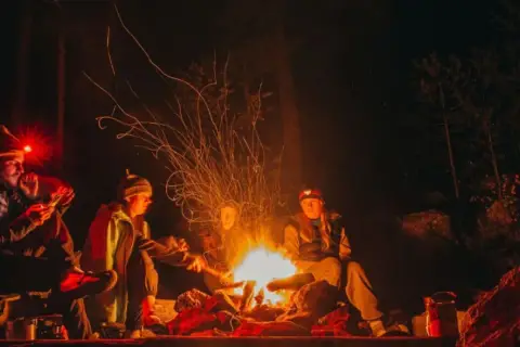 Grupa przyjaciół rozgrzewa się przy ognisku w lesie, zastanawiając się, gdzie na sylwestra w góry. Iskry tańczą w nocnym powietrzu.