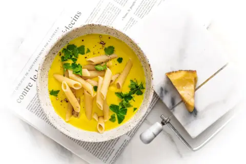 Zupa neapolitańska z makaronem penne i natką pietruszki, podana na gazecie. Obok kawałek sera z ostrzem do sera.