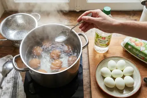 Une main ajoute du vinaigre blanc dans l'eau bouillante où cuisent des œufs. C'est une astuce pour éviter que les œufs durs ne se fendent.