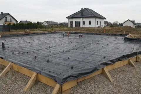 Przygotowane wykopy pod fundamenty, zbrojenie i izolacja przeciwwilgociowa czekają na beton. W tle domy jednorodzinne.