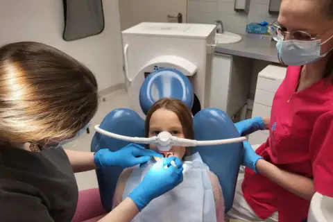 Dziecko u dentysty z maską do podawania gazu rozweselającego. Dwie higienistki dbają o komfort małego pacjenta podczas zabiegu.