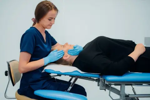 Fizjoterapeuta szczękowy delikatnie masuje twarz pacjentki leżącej na kozetce.